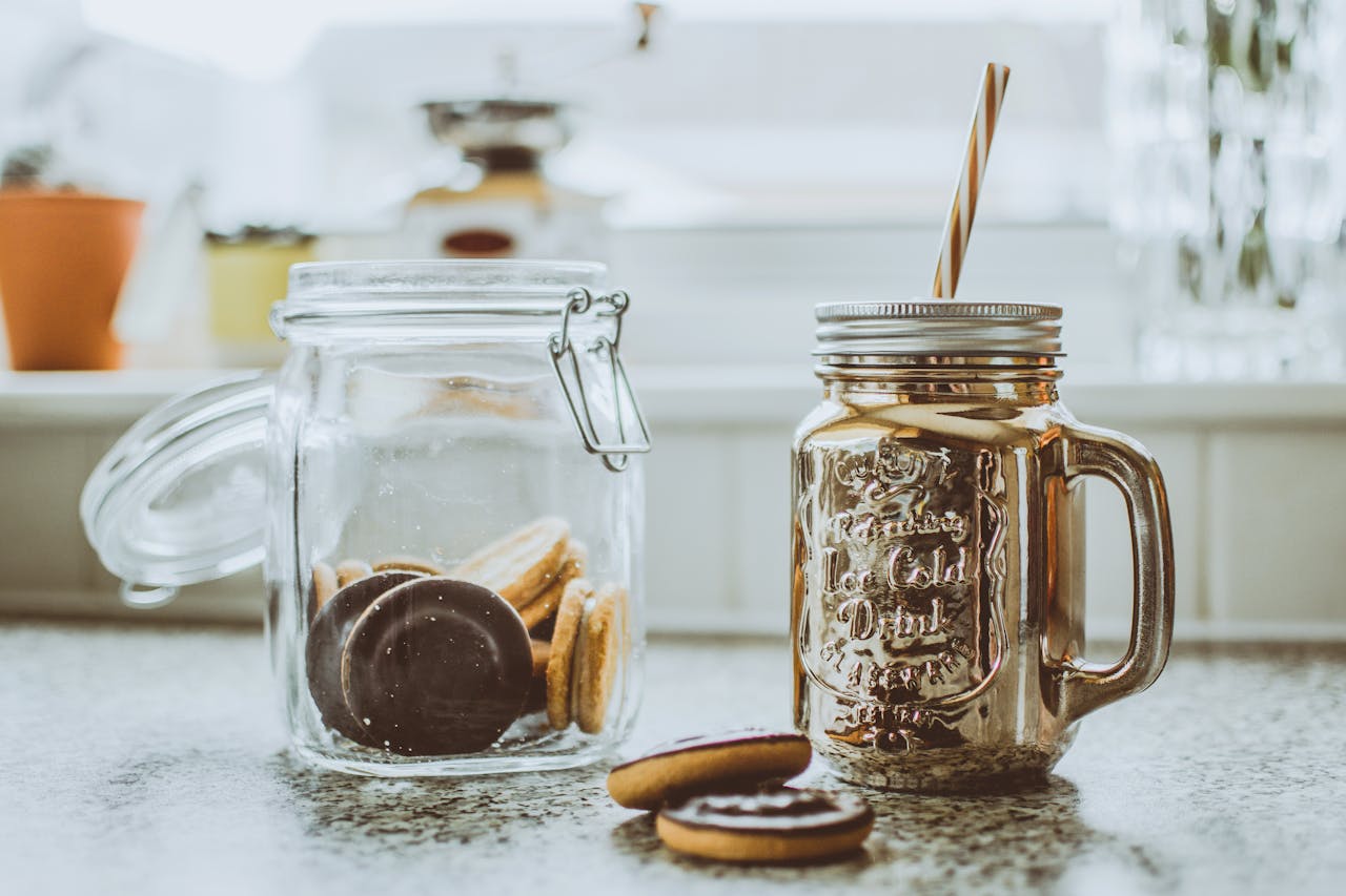 contact-img Glass of milkshake with cookies in a jar on a sunny kitchen countertop.
