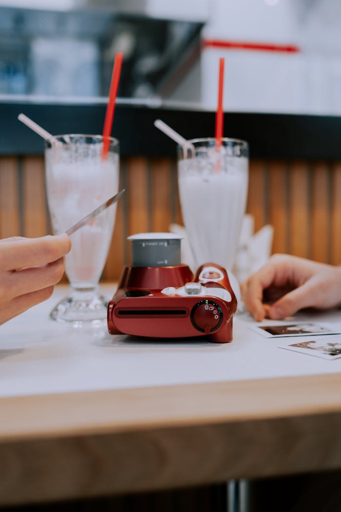 Two milkshakes with straws beside a red camera on a table, hands visible.